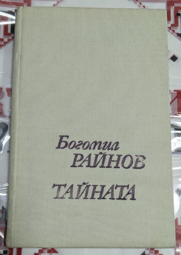 Корица на книгата Тайната от Богомил Райнов, изчистен дизайн с текст в лилав шрифт върху светъл платнен фон.