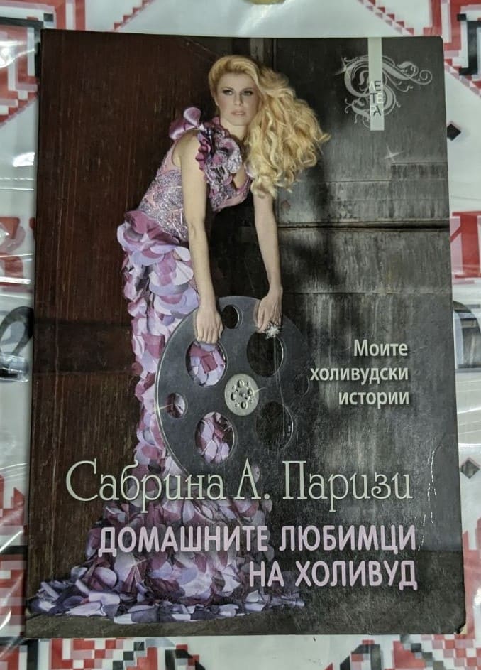 Корицата на книгата „Домашните любимци на Холивуд“ от Сабрина А. Паризи – жена в лилава рокля с филмова лента, символ на Холивуд и блясъка.